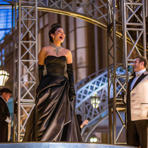 Vanessa Hudgens celebrating 'Gigi' Broadway opening night. She stands radiant, beaming with joy in a stunning black strapless gown with a voluminous skirt and a flowing train. Adorned with a lavish necklace and elegant gloves, she is captured mid-laugh amidst a backdrop of ornate stage decor and dramatic lighting. Surrounding actors in formal attire share in the celebratory moment.
