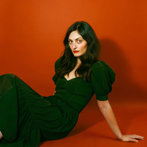 Phoebe Tonkin, in a black and white close-up photo, sitting on a neutral background. She wears a ruffled dress and has long dark wavy hair. Direct gaze.