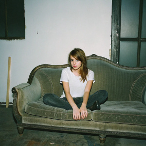 Emma Roberts, sitting on a vintage couch. Casual attire, simple white t-shirt, brown hair loosely styled. Studio lighting, minimal background.