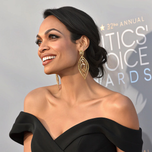 Rosario Dawson at Critics' Choice Awards, wearing black off-shoulder gown. Friendly smile, makeup, and gold earrings complement her appearance. Shot against plain background