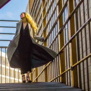 Amber Heard, with her flowing blonde hair, standing alone on a bright, sunny day. She is dressed in a casual outfit, a light herringbone coat paired with a flowing black dress and high heels. Simple, clear sky background