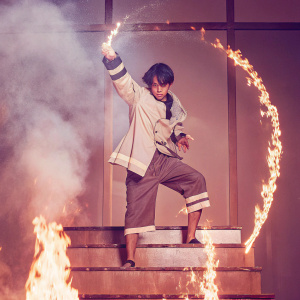 Shinra Kusakabe from Fire Force standing on fiery platforms in a dynamic pose. Dark and smoky background.