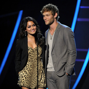Youthful actors Vanessa Hudgens and Alex Pettyfer at an awards show. Vanessa in a leopard print dress and blazer. Alex in a gray suit. Dark background with blue accents.