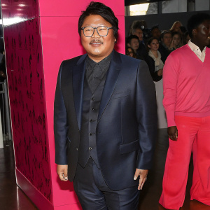 Benedict Wong, dressed in a dark blue suit, glasses perched on nose, smiling warmly. Crowded awards ceremony backdrop