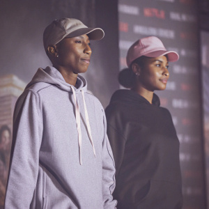 Pharrell Williams and Helen Lasichanh at the 'The Black Godfather' Netflix premiere. They are dressed in casual attire, Pharrell in a blue hoodie and cap, Helen in a black hoodie and pink cap. Against a bright, colorful backdrop featuring Netflix branding.