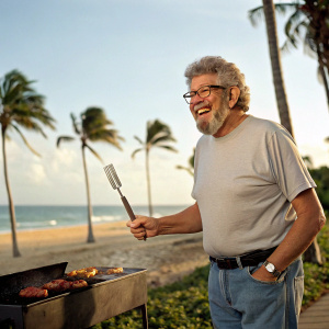 Rolf Harris, with his signature bushy beard and curly hair, smiling widely in a casual t-shirt and jeans at a beachside barbecue.