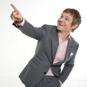 Martin Freeman, dressed in a gray pinstriped suit and light pink shirt, poses with a cheerful smile and a finger pointed forward in a well-lit, plain white studio setting.