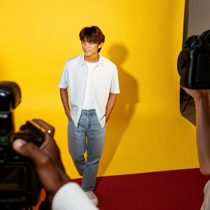 Lee Junho at Cannes Film Festival. Wearing casual attire, white shirt and jeans. Dark background with red carpet and flashing cameras.