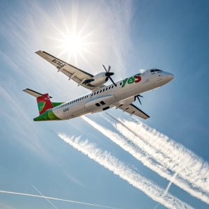 An ATR 72-600 aircraft, operated by TAP Air Portugal, soaring through a bright, clear sky. The plane's white body contrasts with green accents, showcasing its unique tail design. The contrail, a mix of vapor and particles, trails behind it as seen from below.