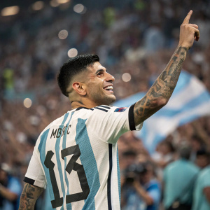 Cristian Romero, wearing Argentina's national team jersey with number 13. Tattoos visible on arm. Cheerful smile. Crowded stadium backdrop.