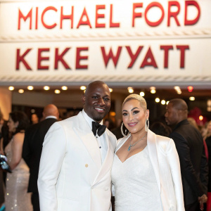 Michael Ford and Keke Wyatt, a black man and white woman, are attending a gala event. Both dressed in elegant white attire. Large white banner with red text behind them. The scene is bustling with guests and fans.