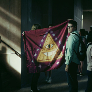 Bill Cipher, character from Gravity Falls, featured on a vibrant red banner. Purple and gold colors dominate the illustration. Event backdrop at a fan convention.