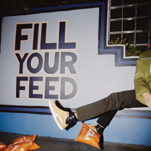 Doritos advertisement, vibrant social media post. Graffiti-styled backdrop. 'FILL YOUR FEED' slogan in bold. Orange and black colors.