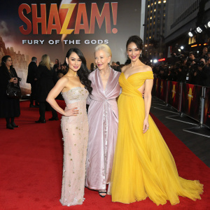Lucy Liu, Helen Mirren, and Rachel Zegler standing together on a bustling red carpet. They pose confidently in front of an enormous 'Shazam! Fury of the Gods' promotional banner.