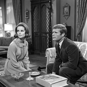 Leigh Taylor-Young and Ryan O'Neal in vintage black and white film scene. Set design reflects 1960s era, with antique furniture and decor.