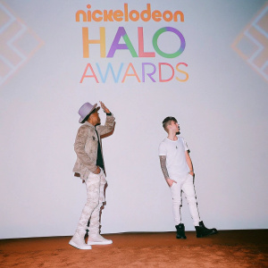 Nick Cannon and Justin Bieber at the Nickelodeon HALO Awards. Both are standing on a stage with a bright, colorful backdrop featuring the event's logo. Nick wears a gray fedora and camouflage jacket, while Justin is in a simple white t-shirt.