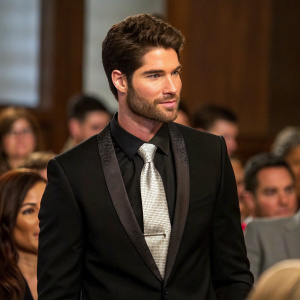 Nick Bateman at a press conference. Black suit, silver tie. Crowded room, blurred faces in background.