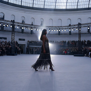 Naomi Campbell on the runway. Wearing a stunning fringe gown at Paris Fashion Week. Crowded auditorium with photographers in the background.