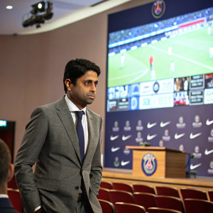 Nasser Al-Khelaifi at a press conference, wearing a grey suit and tie. Behind him is a large screen showing a montage of football matches.