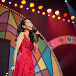 Karen Mok, a vibrant performer, smiles brightly on stage. Multicolored backdrop with shapes, illuminated by warm lights.