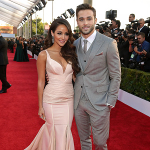 Melanie Iglesias and Ryan Guzman, both Hispanic, at a glamorous award show. Wearing formal attire, Melanie in a stunning pale peach gown, Ryan in a sophisticated gray suit. Surrounded by flashing cameras on the vibrant red carpet.