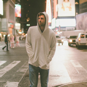 Nomar Garciaparra, wearing a light grey hoodie and jeans, standing in the middle of Times Square at night. Bright lights and billboards all around.