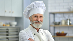 Caucasian middle aged male chef in a chef's hat with arms crossed wears apron standing in restaurant kitchen and smiling