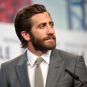 Jake Gyllenhaal, actor with beard and styled hair, wearing gray suit, crisp white shirt, and gray tie. Photo taken at press conference. Background is blurred, focusing on Jake.