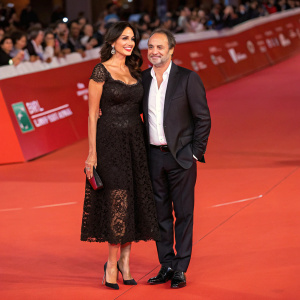 Maria Grazia Cucinotta and Giulio Violati, dressed elegantly, on the vibrant red carpet. Maria in a stunning black lace dress with matching high heels. Giulio in a tailored dark suit with a crisp white shirt. They pose confidently at the 'Obra' premiere during the 9th Rome Film Festival.