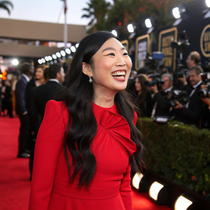Awkwafina, wearing a vibrant red dress, beams brightly on the Golden Globe red carpet. The background buzzes with flashing cameras and eager crowds.