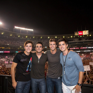 Kendall Schmidt, Carlos Pena Jr., James Maslow, and Logan Henderson from Big Time Rush at a concert. Wearing casual t-shirts and jeans. Stadium lights and fans in background