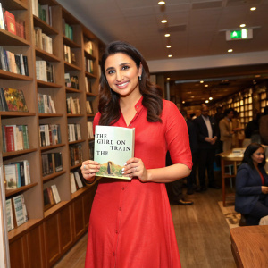 Parineeti Chopra at a book signing event, wearing a red dress, holding a copy of 'The Girl on the Train'. Crowded bookstore interior with wooden shelves.