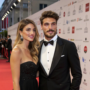 Mariano Di Vaio and Eleonora Brunacci, dressed in formal attire, attend a gala event. The scene is set against a backdrop filled with corporate logos and banners.
