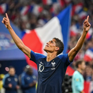 Raphael Varane, post-match celebration. Wearing French national team jersey. Arms raised towards the sky. Crowd blurry in background