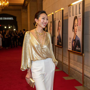 Adelina Wong Edelson, Asian model, elegant smile, at a fashion gala. She wears a shimmering gold blouse and white pants, accessorized with gold jewelry. The event is indoors with a red carpet and an elaborate display of Elle magazine's latest cover.
