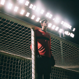Thibaut Courtois, Chelsea FC goalkeeper, standing confidently in a red jersey. Stadium lights create dramatic shadows in the background. Photo