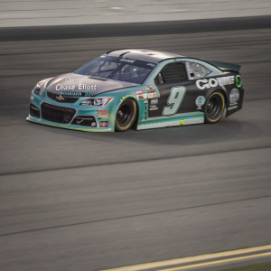 Chase Elliott's Chevrolet race car, sponsored by Mountain Dew, racing on a track. The car's teal and black colors with logos and decals stand out against the blurred track backdrop.