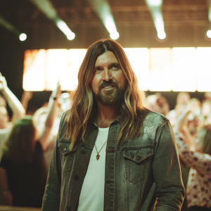 Billy Ray Cyrus, long blonde hair and beard, wearing a denim jacket, poses in a bustling concert venue.