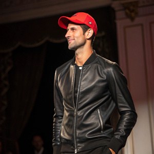 Novak Djokovic at a charity event. Wearing a black leather jacket and a red cap. Dark, elegant background