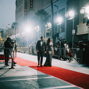 Serj Tankian and Angela Madatyan walking on a Hollywood Boulevard red carpet. Behind them, bright lights and paparazzi flash