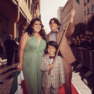 Angelica Masiel Padron, Angelica Vale, and Daniel Nicolas Padron Vale posing for photos at a star-studded event on Hollywood Boulevard. Angelica Masiel Padron wears a vibrant green dress. Daniel Nicolas Padron Vale, a young boy, in a plaid outfit, holds a grand Mexican flag. Crowded street, festive atmosphere