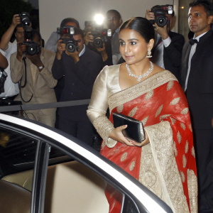 Vidya Balan at the Cannes Film Festival. Wearing a vibrant red saree with intricate designs. Pearl necklace. Surrounded by flashing cameras and a crowd.