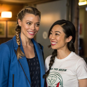 Lauren German and Aimee Garcia on the set of a TV show. Lauren is wearing a blue jacket and has a braid. Aimee is in a white t-shirt with graphic design. Both are smiling