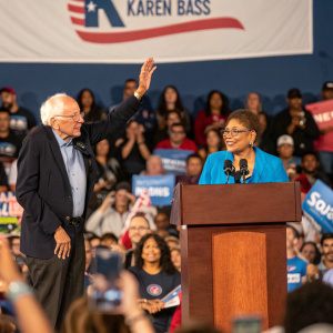 Bernie Sanders and Karen Bass addressing a crowd at a bustling political rally. The backdrop features a banner with Karen Bass's campaign logo.