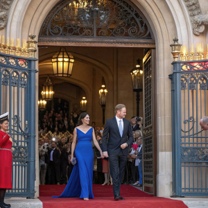 Meghan Markle and Prince Harry exiting a grand palace. She is adorned in a royal blue evening gown. Crowds gathered