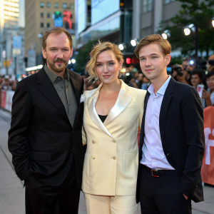 Ralph Fiennes, Kate Winslet, and David Kross attending a press conference for a new film. Fiennes in a black suit, Winslet in a cream-colored blazer, Kross in a casual shirt. The backdrop is a bustling film festival street with fans and paparazzi.