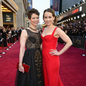 Thea Sharrock and Emilia Clarke at the Oscars. Thea in a black dress with intricate details and matching clutch. Emilia in a vibrant red gown. They pose on the bustling red carpet. Crowds and barricades visible in the background. The scene captures the excitement of a star-studded gala.