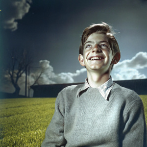 Andrew Garfield, young actor, wearing a grey sweater, standing on a vibrant green grass field with a cheerful smile.