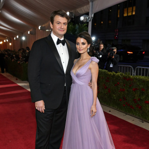 Nathan Fillion and Mikaela Hoover, both dressed formally, attend the Met Gala. Nathan in a black tuxedo, Mikaela in a flowing lavender gown. Red carpet event.