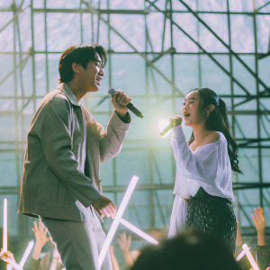 So Ji-sub and Cho Eun-jung attending a vibrant K-pop concert. The stage is lit with neon lights, showcasing a dynamic performance.